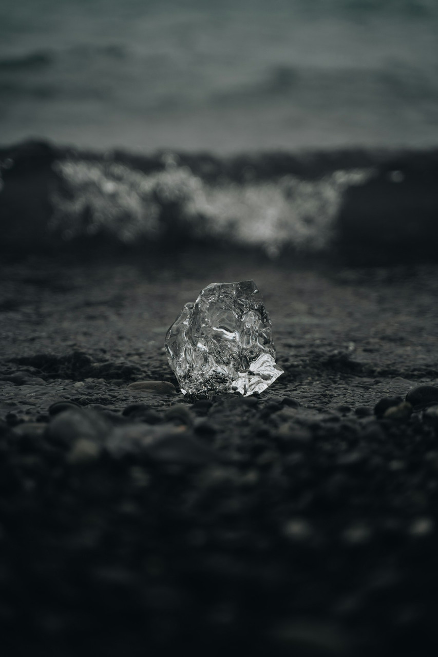 A clear chunk of ice on a dark pebble beach near the ocean, Ultra