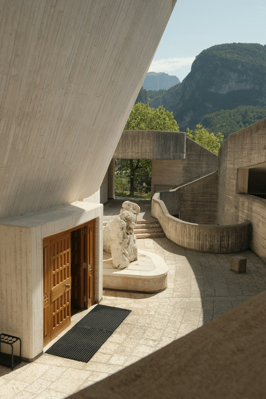 A concrete courtyard with wooden doors and a stone sculpture beneath mountain peaks, Ultra