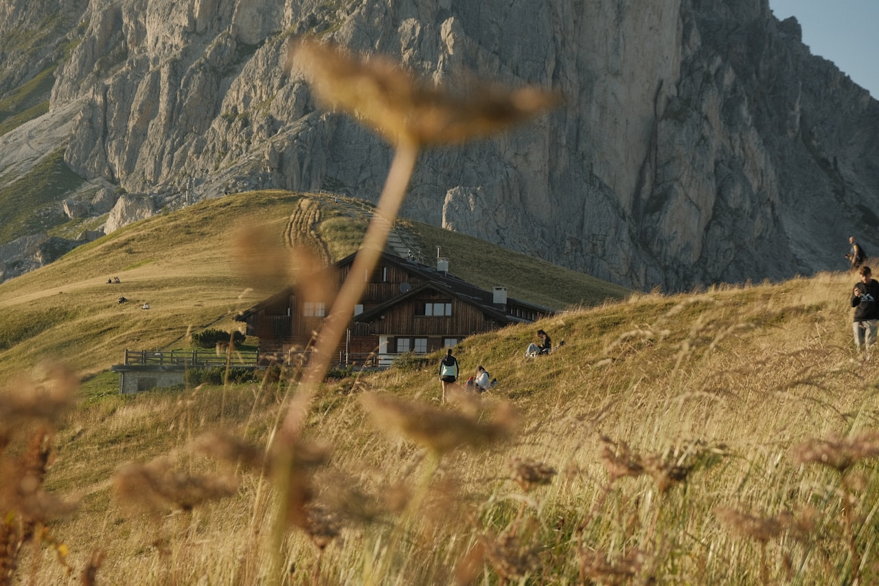 People on a grassy hillside with a wooden mountain hut below rocky peaks, Ultra
