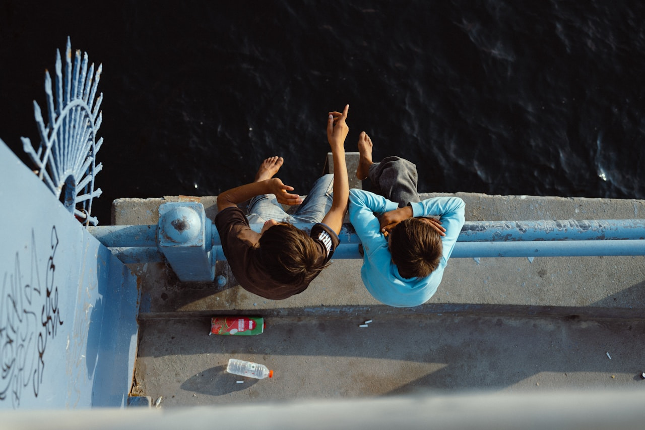 Two people sitting on a blue bridge railing pointing toward dark water below, Ultra