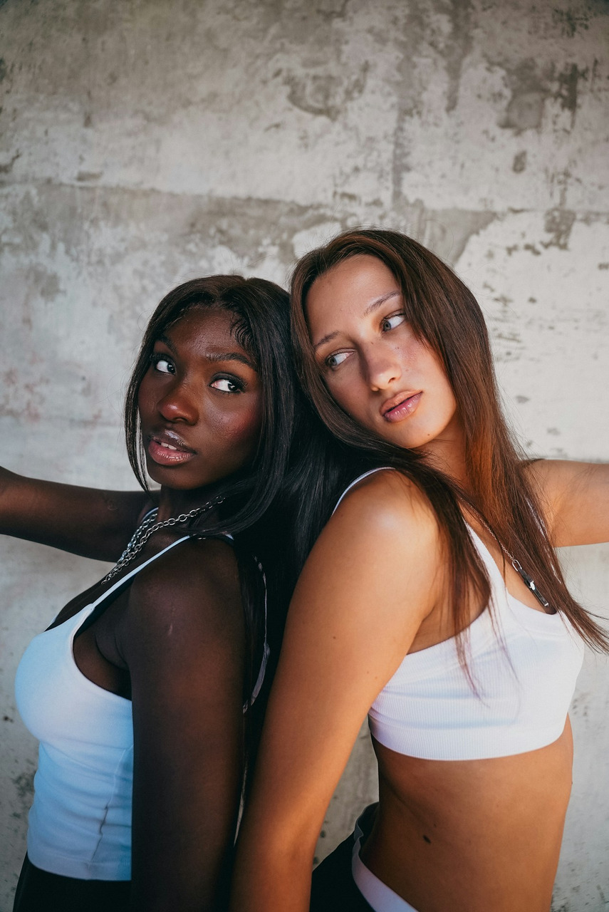 Two women in white crop tops standing back to back against a concrete wall, Ultra