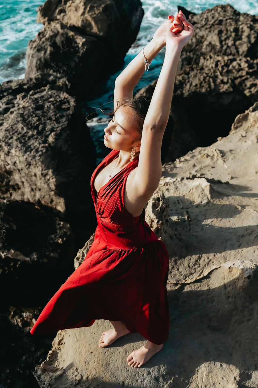 Woman in red dress with raised arms on rocky coast, Ultra